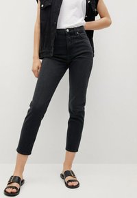 Jeans negros de cintura alta con un corte ajustado, hechos de mezclilla, con una textura suave y dos bolsillos frontales. Llevados con sandalias negras de plataforma.