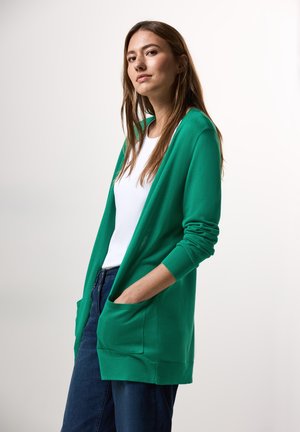 Cardigan vert à manches longues, à ouverture devant, avec deux poches frontales, superposé sur une chemise blanche, porté avec un jean foncé. Texture lisse.