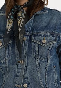 Jeansjacka i medelblått med två bröstfickor, metallknappar och en svart blommig silkescarf knuten runt halsen.