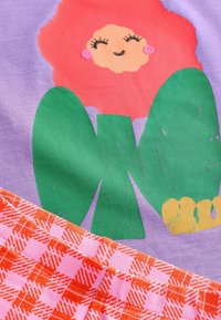 Lilla stof prydes af en smilende blomsterkarakter med en rød, tekstureret kant, parret med orange og pink ternede detaljer nedenfor.