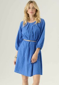 Abito azzurro a pois con maniche a sbuffo e vita cinchata, dotato di una cintura beige e realizzato in un tessuto morbido e leggero.
