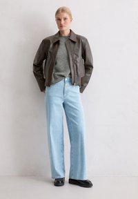 Bruine leren jas met ritssluiting, grijze trui en lichtblauwe wijde jeans. Zwarte schoenen maken de outfit compleet. Effen achtergrond.