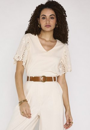 Donna con capelli ricci che indossa un top color crema con scollo a V e maniche di pizzo, pantaloni crema a vita alta, cintura marrone e gioielli in oro.