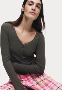 Maglietta a maniche lunghe grigio scuro con scollatura a V ampia, dettagli con bottoni e cuciture dorate, abbinata a pantaloni della pigiama a quadri rosa.