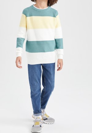 Kind trägt einen blau-gelb-weiß gestreiften Pullover, blaue Jeans und graue Turnschuhe mit gelben Schnürsenkeln, steht vor weißem Hintergrund.