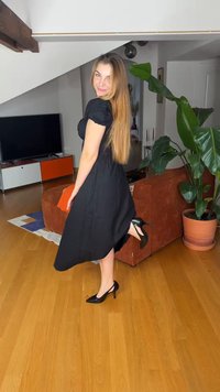 Γυναίκα με μακριά μαλλιά που φοράει μαύρο φόρεμα και ψηλοτάκουνα, ποζάροντας σε εσωτερικό χώρο κοντά σε καφέ καναπέ και μεγάλο γλαστράκι σε σαλόνι.