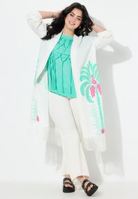 Valkoinen, fringed-kimono, jossa on vaaleanpunaisia ja vihreitä palmukuoseja, päällä teal-välinen toppi, yhdistettynä valkoisiin leveälahkeisiin housuihin ja mustiin korkokenkiin.