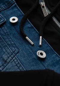 Giacca di jeans con un hoodie nero, caratterizzata da hardware argentato, una cerniera e lacci neri con estremità metalliche.