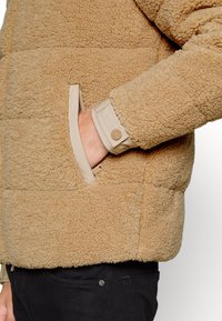 Tanskleurige fleecejas met een gestructureerd oppervlak, ritszakken en een lichte mouw met een knoopdetail.