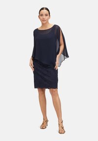 Námořnický chiffonový plášť přes krajkovou tužkovou sukni se zakulaceným lemem. Modelka nosí sandály na klínku se zlatými pásky a pózuje na bílém pozadí.