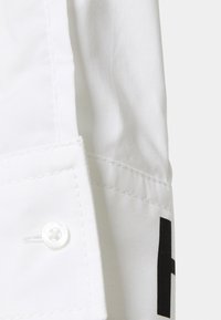 Gros plan d’un poignet en tissu blanc avec un bouton blanc et des coutures visibles, partie d’un vêtement avec une impression partielle en lettre noire.