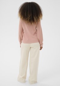 Femme aux cheveux bouclés portant un haut à manches longues rose et un pantalon large crème, se tenant de dos sur un fond blanc.