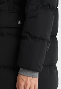 Manga de chaqueta de plumas negra con textura acolchada, tela suave y un botón de presión plateado. Un puño gris asoma por debajo de la manga.
