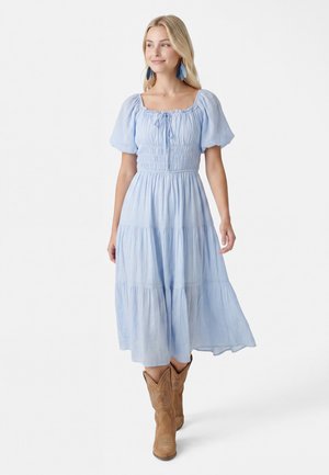 Kvinde iført en lyseblå midikjole med puffede ærmer, smocktalje og beige cowboystøvler, stående foran en ensfarvet hvid baggrund.