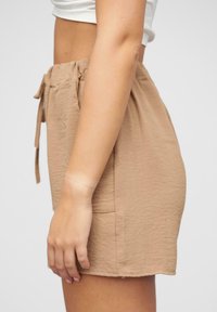 Beige šortky vyrobené z texturované tkaniny, s pasem na šňůrku a bočními kapsami. Spodní lem je hladký, s minimalistickými designovými detaily.