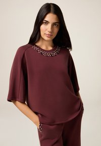 Bordeaux sweatshirt met korte, brede mouwen, versierd met glanzende zilveren en donkere edelstenen rond de halslijn en zakken, gemaakt van een zachte stof.
