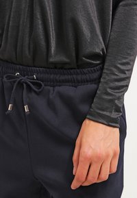 Dark blue joggers with an elastic waistband, adjustable drawstring, and silver-tone hardware. Black, shiny long-sleeve top with textured fabric.