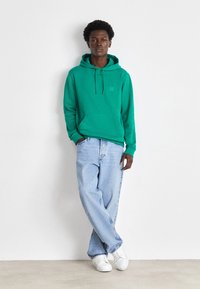 Green hoodie made of soft fabric with a front pocket, paired with light blue baggy jeans and white sneakers, standing against a light background.