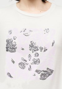 Λευκό μπλουζάκι με ασημένια παγιέτα σε σχέδια λουλουδιών και φύλλων στο στήθος, φορεμένο από ένα άτομο με κοντά σκούρα μαλλιά.