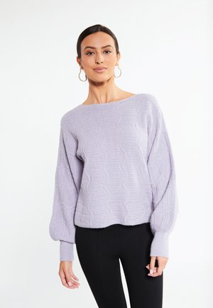 Frau mit dunklem Haar, die große Creolen-Ohrringe, einen hellvioletten strukturierten Pullover und schwarze Hosen trägt, steht vor einem weißen Hintergrund.