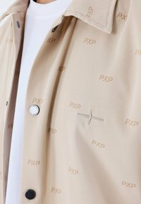 Chaqueta beige con un patrón repetido de "PXP", botones de presión plateados y acentos bordados. Presenta una textura suave y un diseño de cuello clásico.