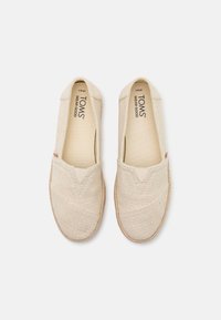 Paire de chaussures TOMS beige tissées sans lacets, à bouts arrondis, avec "TOMS WEAR GOOD" imprimé à l'intérieur, sur fond blanc.
