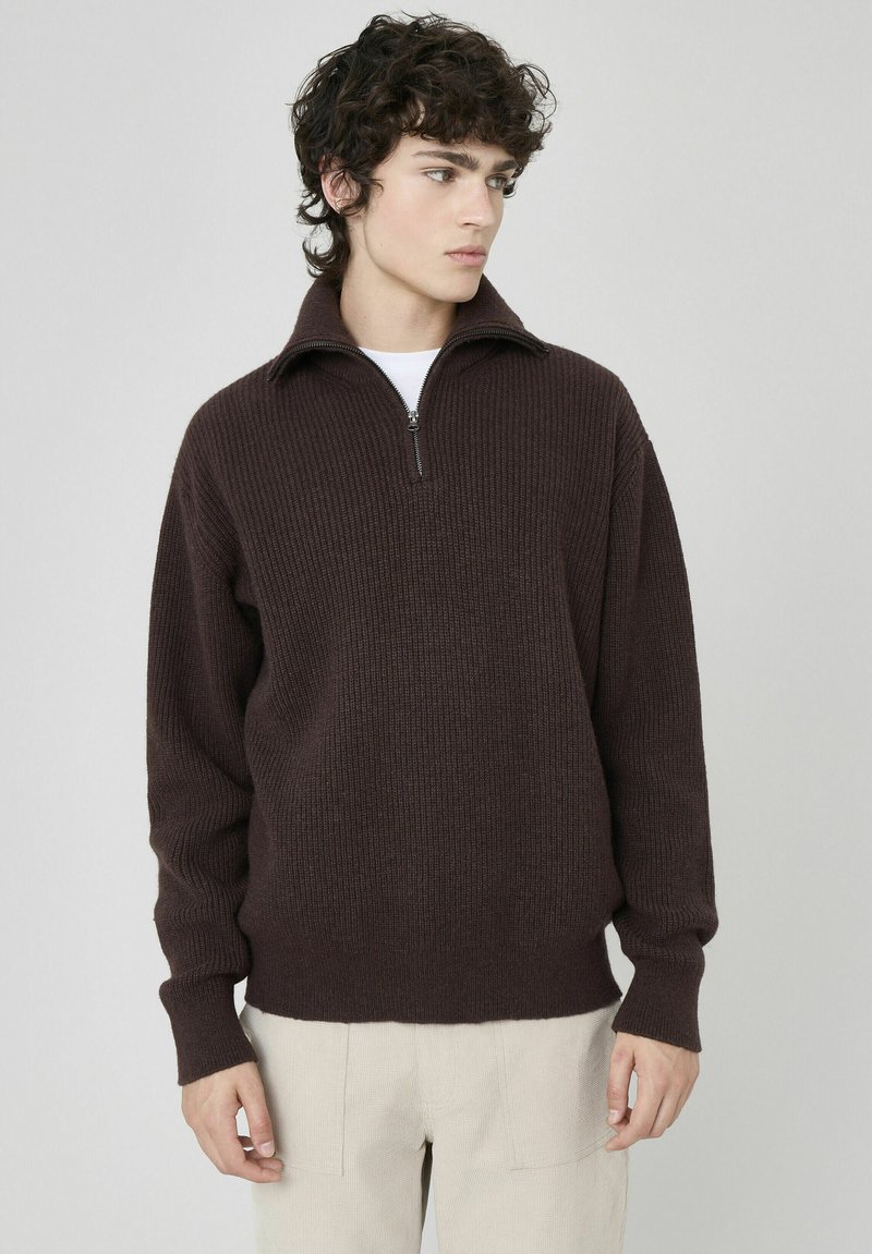 Pull en maille texturée marron avec un col zippé quart et un design en côtes. Porté avec un pantalon de couleur claire, style décontracté.