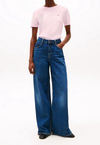 Světle růžové tričko s krátkým rukávem a logem, spojené s vysokým pasem a širokými modrými džíny. Černé boty dotvářejí celý outfit.
