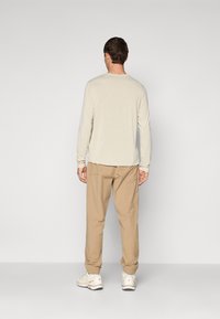 Man standing facing away wearing a beige long-sleeve shirt, light brown jogger pants, and white sneakers on a plain background.