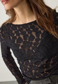 Haut à manches longues en dentelle noire avec motifs floraux, encolure bateau et texture semi-transparente, associé à un tissu gris texturé en dessous.