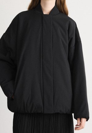 Frau trägt eine übergroße schwarze Steppjacke mit hohem Kragen und elastischem Saum, kombiniert mit einem plissierten schwarzen Rock vor neutralem Hintergrund.