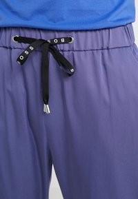 Luźne fioletowe spodnie z elastycznym pasem, z czarnym sznurkiem do regulacji z logo oraz srebrnymi końcówkami. Gładka tekstura.