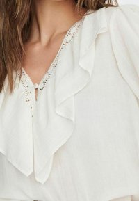 Weiße Bluse mit tiefem V-Ausschnitt und Spitzenbesatz sowie geschichteten Rüschendetails am Ausschnitt, aus weichem, leichtem Stoff mit fließender Textur.