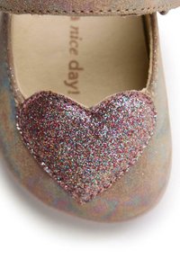 Chaussure beige avec un accent en forme de coeur rose pailleté. Matière douce avec une surface texturée, bordée d'une couture autour du coeur.