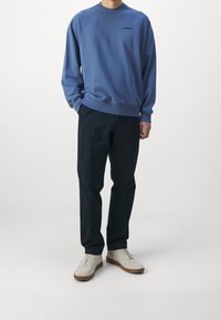 Blå crewneck-tröja med ribbade muddar och fåll, med en liten svart logotyp. Matchas med mörka byxor och ljust färgade sneakers.