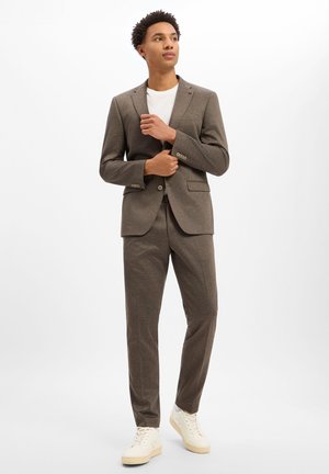 Young man standing wearing a brown suit with a white shirt and cream sneakers, adjusting his jacket in a plain white background.