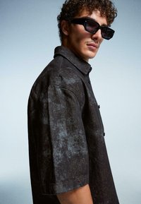 Uomo con capelli ricci che indossa occhiali da sole neri e una camicia a maniche corte button-up di un colore scuro, posando su uno sfondo semplice.