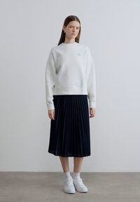 Sweat-shirt blanc avec logo vert, accompagné d'une jupe plissée noire et de baskets blanches. Coupe ample, tissu texturé, jupe longueur genoux.