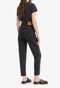 Jean en denim noir taille haute avec des jambes fuselées, arborant une étiquette en cuir au dos, et portés avec des chaussures glissantes noires.