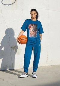 Sieviete zilā sporta apģērbā, turot oranžu basketbola bumbu, stāvot pie plaisātas baltas sienas ar ēnu un basketbola grozu virs galvas.