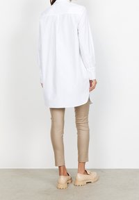 Camicia bianca a maniche lunghe con colletto, taglio dritto e polsini abbottonati, abbinata a pantaloni beige attillati e scarpe chunky di colore chiaro.