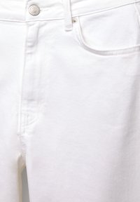 Jean en denim blanc en gros plan montrant la poche avant, la passant de ceinture, le bouton argenté et les détails de rivets.