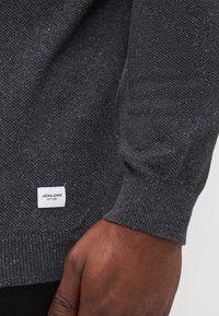 Dark gray knitted sweater with a textured pattern, ribbed cuffs, and a small white brand label on the left side.