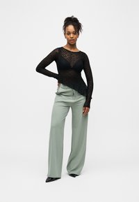 Moteris stovinti, viena ranka atsikišusi į klubą, vilkinti juodą permatomą ilgomis rankovėmis nėriniuotą viršutinę dalį, žalius plačius kelnių kojų šonus ir juodus smailius batus.