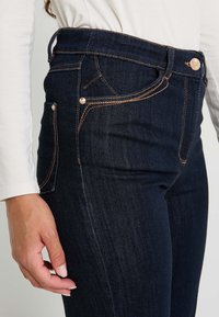 Jeans en denim bleu foncé à taille haute, avec des finitions en métal cuivré, des coutures double piqué et un design de poche sur le côté.
