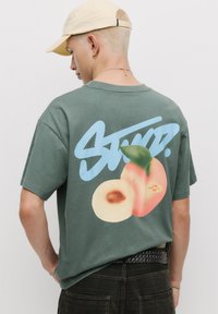 Grünes Baumwoll-T-Shirt mit einem großen Grafikdruck auf der Rückseite, der eine Pfirsichabbildung und das Wort "STUFD." in Blau zeigt. Kurze Ärmel, lockere Passform.