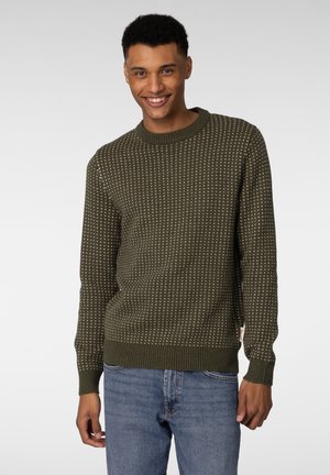 Mørkegrønn strikket genser med et teksturert mønster og ribbede mansjetter og kant. Båret med lyseblå jeans, med en rund halslinning.