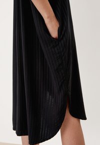 Czarna sukienka w pionowe paski, z kieszeniami po bokach i lekko zaokrąglonym dołem. Wykonana z miękkiego materiału o gładkiej teksturze.