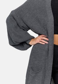 Cardigan grigio a maglia con maniche oversize, aperto davanti e due tasche. La texture a coste aggiunge profondità al design.