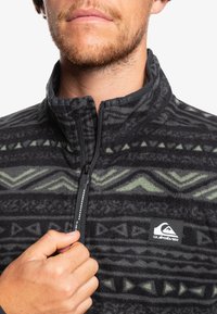 Svart fleecejacka med gröna geometriska mönster, halvdragkedja, hög krage och en Quiksilver-logotyp på vänster bröst.